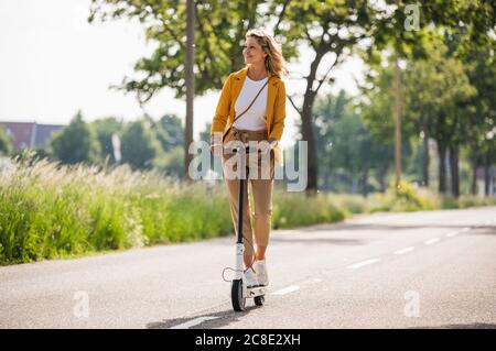 Mature woman riding electric push scooter on road during sunny day Stock Photo