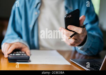 Senior man holding smart phone while using calculator and laptop at home Stock Photo