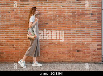Young woman carrying groceries in mesh bag while walking on footpath by brick wall Stock Photo