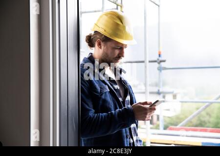 Construction worker wearing helmet using smart phone by window at constructing house Stock Photo
