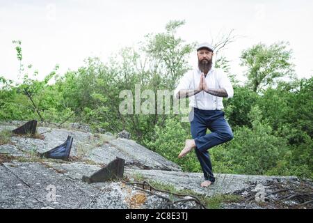 Bearded man wearing cap practicing yoga on rock against trees Stock Photo