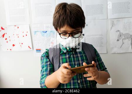 Boy wearing mask using mobile phone while standing against wall in school Stock Photo