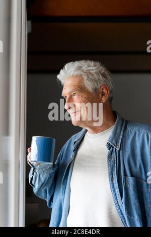 Thoughtful senior man smiling while drinking coffee by window at home Stock Photo