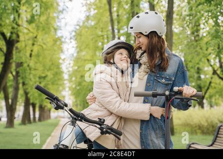 Girl with bicycle embracing mother while standing against trees in city park Stock Photo