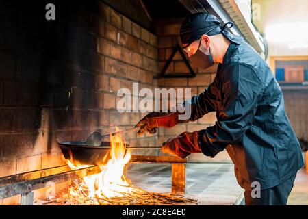 Traditional cooking of paella in restaurant kitchen, chef wearing protective mask Stock Photo