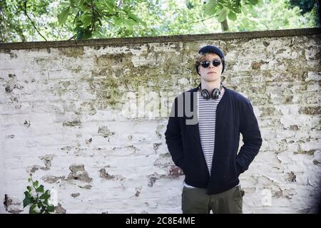 Portrait of young man with headphones wearing sunglasses and cap in front of weathered wall Stock Photo