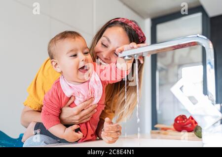 Close-up of smiling mother holding cute baby girl playing with faucet in kitchen Stock Photo