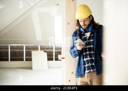 Construction worker wearing helmet using smart phone while standing by wood in renovating house Stock Photo