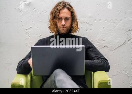 Bearded young man using laptop while sitting on armchair against wall at home Stock Photo