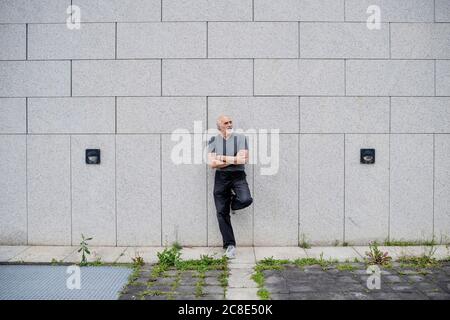 Thoughtful senior man with arms crossed leaning on wall in city Stock Photo