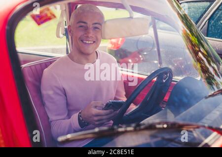 Portrait of smiling teenage boy sitting in vintage car with smartphone looking out of window