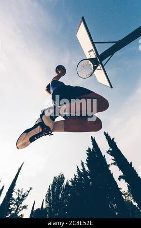 Directly below view of teenage player dunking ball in basketball hoop against sky Stock Photo