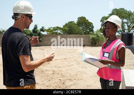 Coworkers wearing helmets discussing while standing at construction site during sunny day Stock Photo