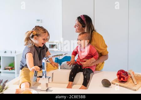 Mother and cheerful daughters playing with water in kitchen sink at home Stock Photo