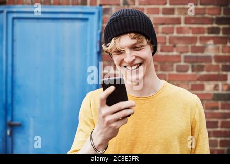 Portrait of happy young man wearing cap looking at smartphone in front of brick wall Stock Photo