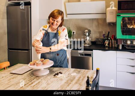 Smiling mature woman icing cupcakes on cakestand in workshop Stock Photo