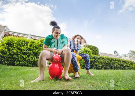 Happy siblings playing with hoppity horses on grassy land against sky in yard Stock Photo