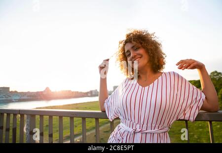 Smiling mid adult woman with curly hair leaning on railing against sky at sunset Stock Photo