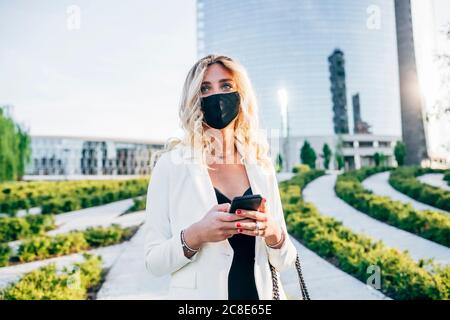 Businesswoman wearing face mask using smart phone while standing in city Stock Photo