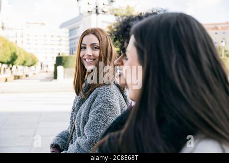 Smiling young woman looking at female friends while walking in town square Stock Photo