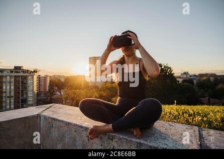 Sporty woman using VR glasses while sitting on retaining wall in city during sunset Stock Photo