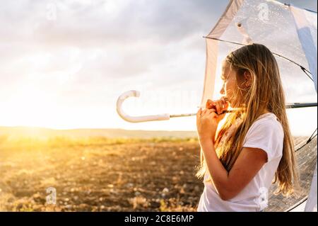 Girl with long blond hair holding umbrella while looking away during sunset Stock Photo