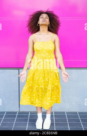 Young woman wearing yellow dress meditating while standing against pink wall