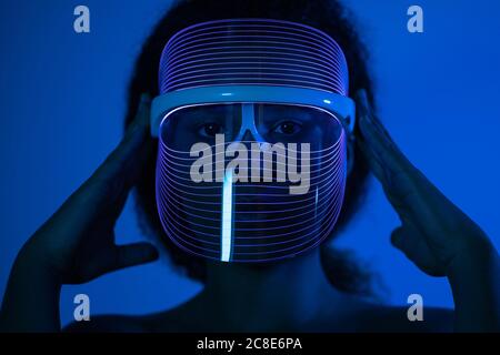 Close-up of woman wearing blue led mask against wall at home Stock Photo