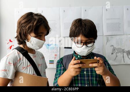 Boy wearing mask looking at friend using smart phone while standing against wall in school Stock Photo