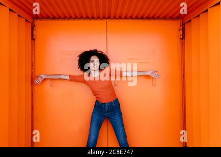 Smiling young woman standing with arms outstretched against orange metal door