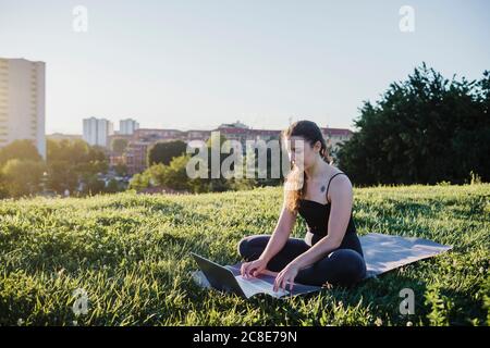 Sporty young woman using laptop while sitting on exercise mat at city park Stock Photo