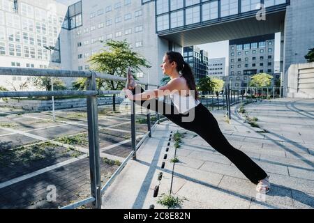 Young sporty woman stretching legs on railing in city during sunny day Stock Photo