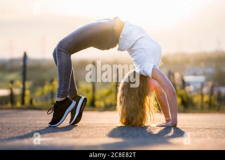 Young flexible woman doing backward bend on road during sunny day Stock Photo