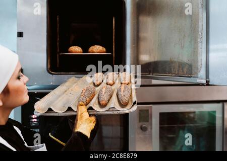 Female baker inserting wholegrain bread on baking pan in oven at bakery Stock Photo