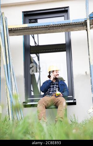Construction worker talking over mobile phone while sitting against window Stock Photo