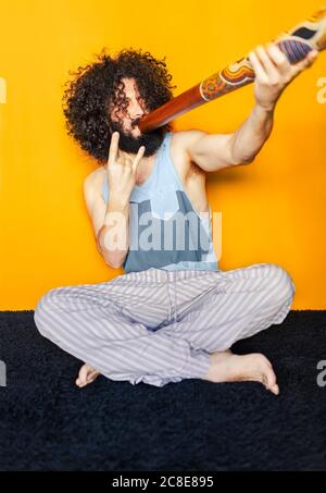 Man with curly hair playing didgeridoo gesturing while sitting against yellow background Stock Photo