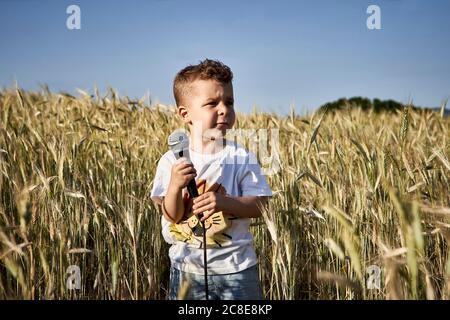 Boy holding microphone looking away while standing amidst crops against clear sky Stock Photo