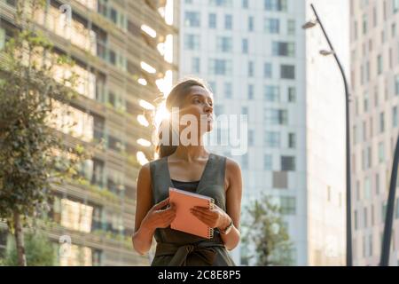 Thoughtful female entrepreneur holding note pad while standing against buildings in modern city Stock Photo