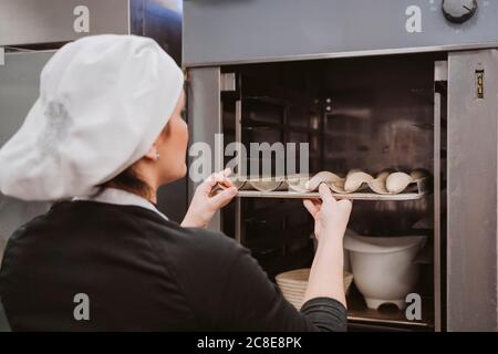 Female baker inserting baking pan in oven at bakery Stock Photo