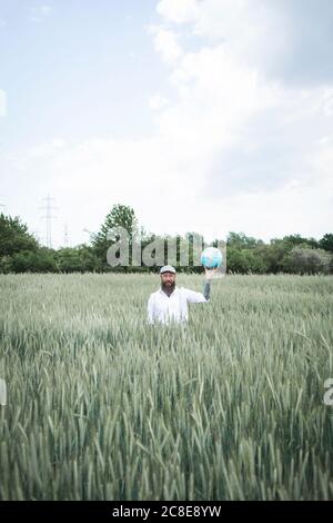 Mature man holding globe while standing amidst cornfield against sky Stock Photo