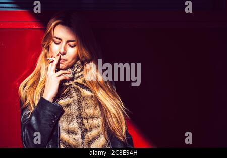 Stylish businesswoman smoking while standing against wall in city Stock Photo