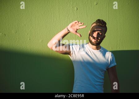 Handsome young man shielding eyes while standing against green wall