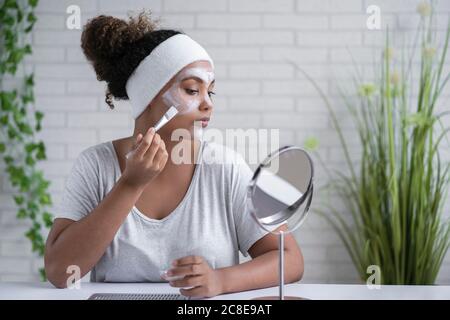 Woman wearing headband applying facial mask while looking in mirror at home