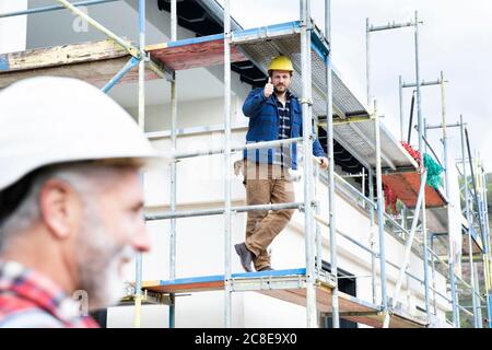 Construction worker showing thumbs up to coworker while sitting on scaffold at site Stock Photo