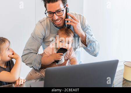 Father talking over smart phone while taking care of daughters at home Stock Photo