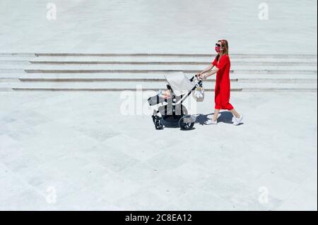 Mother wearing mask pushing son in baby carriage while walking on town square Stock Photo