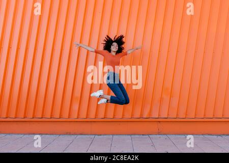 Happy young woman jumping with arms outstretched against orange wall Stock Photo