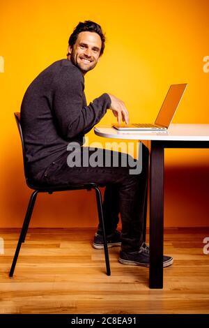 Smiling handsome man using laptop while sitting at table against yellow background Stock Photo