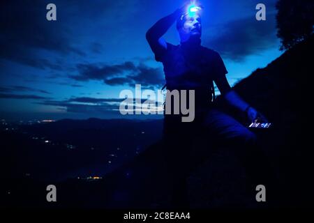 Male hiker with headlamp looking up while standing on mountain at night, Orobie, Lecco, Italy Stock Photo