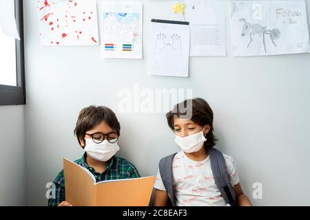 Boys wearing masks reading book while sitting against wall in school Stock Photo